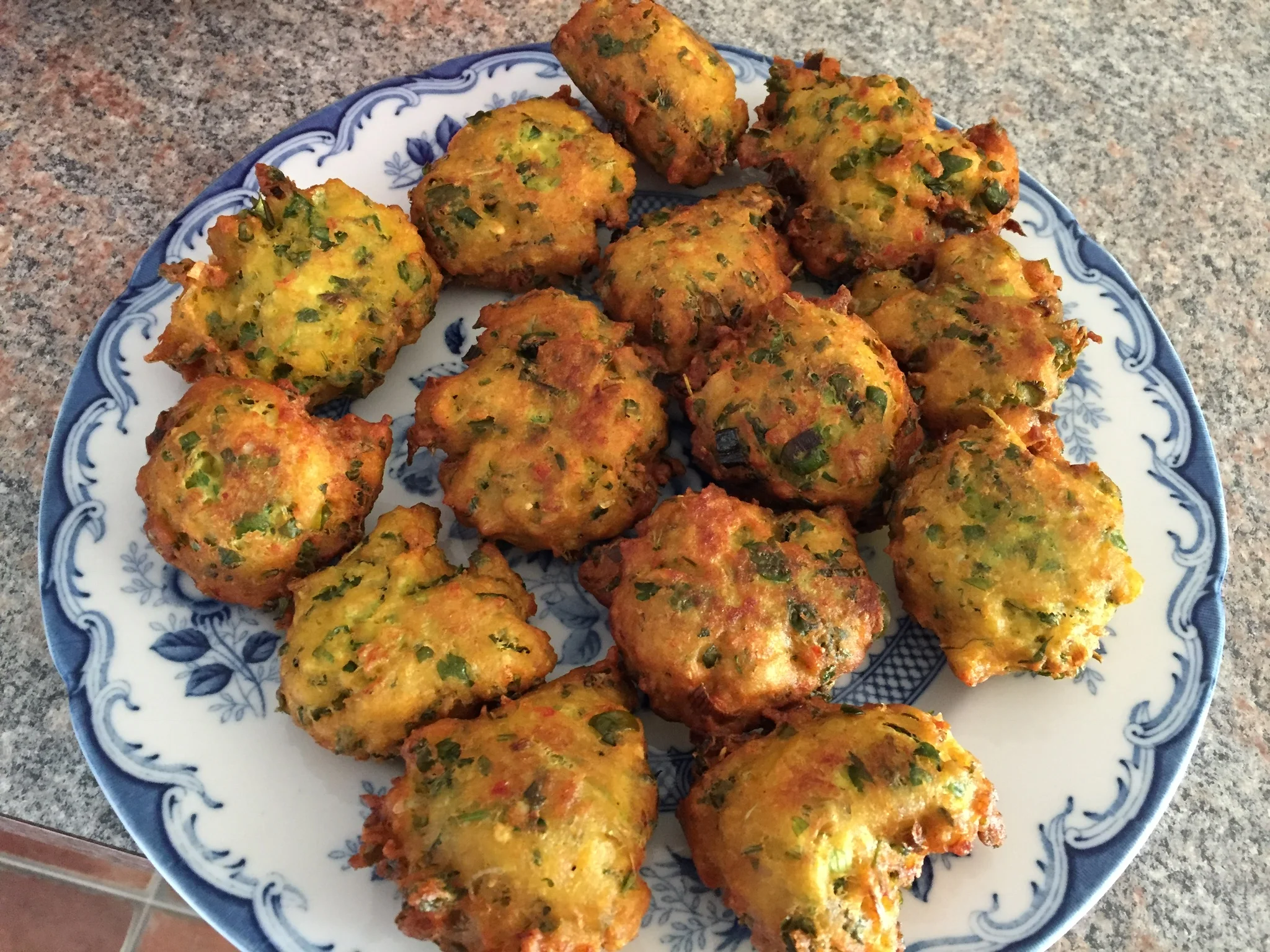 Golden-brown fish fritters speckled with vibrant green herbs and scallions, piled high on a decorative blue-rimmed white plate atop a gray granite surface.